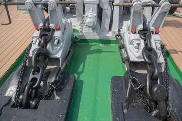 Pair of stationary anchor stoppers mounted on green pedestal on brown wooden deck of vessel with massive black anchor chains attached to them with help of rigging brackets and lanyards.