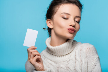 Brunette woman in cozy sweater blowing air kiss and holding blank card isolated on blue.