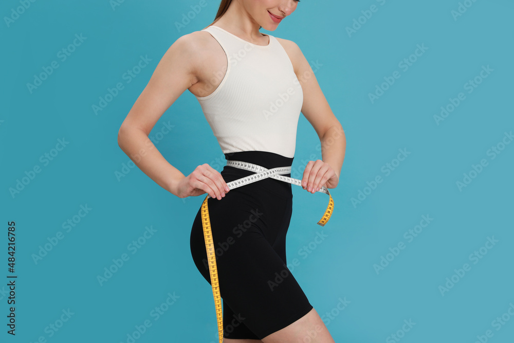 Poster Happy woman in sportswear measuring waist with tape on light blue background, closeup