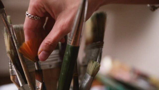 Choosing a brush for painting. Motion shot. Closeup view of young female artist hand grabbing a paint brush from a container with many paintbrushes of different sizes and styles.