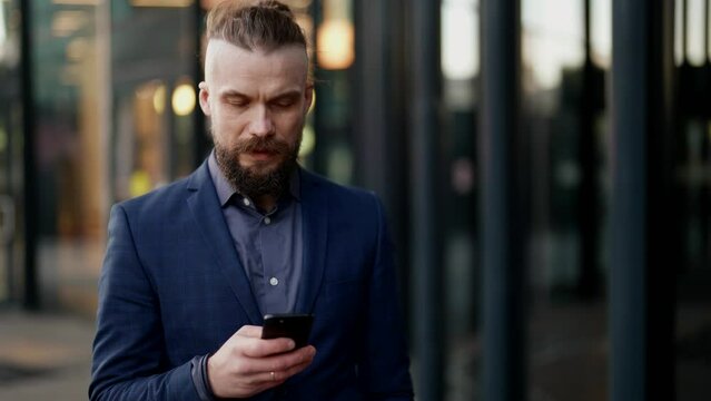 a mustachioed and bearded man in a plaid jacket looks discontentedly at the phone screen and walks along a blurry modern building