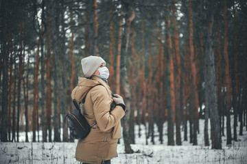 A young girl in winter clothes and in a medical mask is walking in the forest.