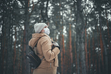 Fototapeta na wymiar A young girl with blond hair in a medical mask walks in the winter forest.