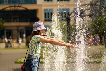 A girl (a teenager) with a red bag, in blue jeans, a striped T-shirt and a summer hat, on a summer alley, laughs and wets her hands in the spray of the fountain. Banner, cover, flyer, layout design