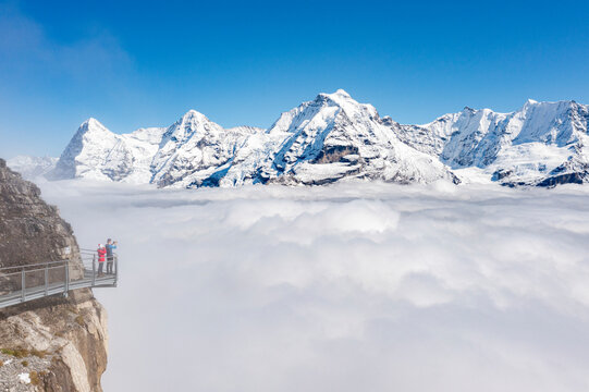 Two people photographing snowcapped Eiger and Monch peaks in fog from elevated walkway, Murren Birg, Bern canton, Swiss Alps, Switzerland