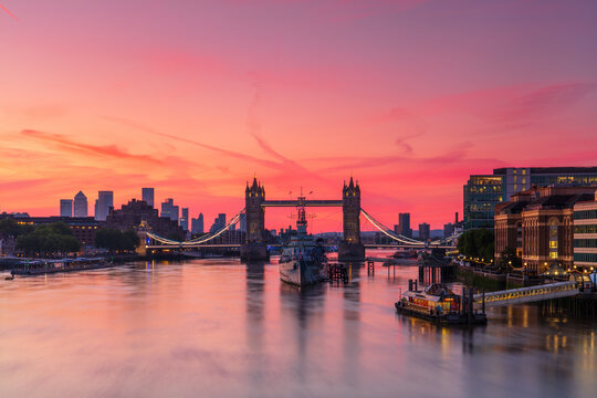 Tower Bridge, River Thames and HMS Belfast at sunrise with pink sky, and Canary Wharf in background, London, England
