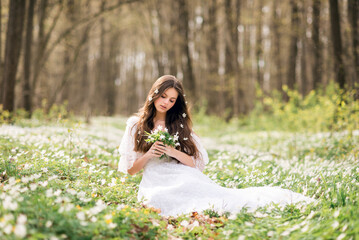 young beautiful woman in a white dress collects primroses. A girl in the spring forest. A bouquet of white anemones in her hands