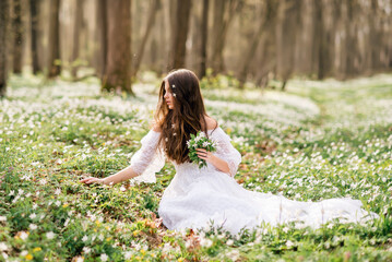 young beautiful woman in a white dress collects primroses. A girl in the spring forest. A bouquet of white anemones in her hands