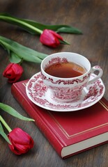 Fototapeta na wymiar reading over tea. Tea in a beautiful tea pair English porcelain, red book and red tulips on a dark background. Soft focus