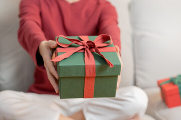 Christmas eve festival season day, hands of young woman, girl holding green gift box with red ribbon, bow on sofa. Happy to get received present on merry xmas. Celebration on New Year festival people.