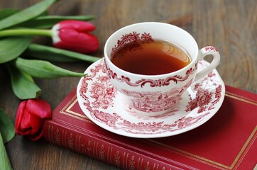 reading over tea. Tea in a beautiful tea pair English porcelain, red book  and red tulips on a dark background. Soft focus