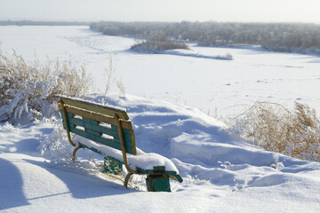 A bench in snowdrifts on a steep bank of a river against the backdrop of a distant forest.