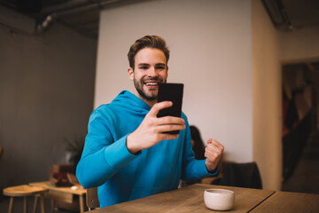 Joyful male blogger reading email with good news about online win in sports betting, cheerful hipster guy enjoying web messaging via cellular technology browsing social networks on smartphone