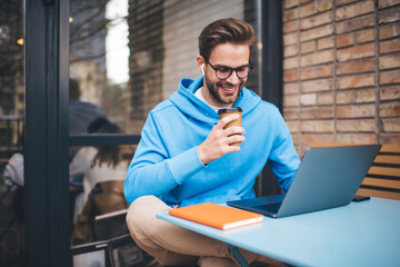 Smiling hipster guy in optical eyewear holding coffee to go enjoying time for doing distance...