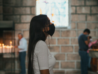 latina girl in profile kneeling praying inside the church she is with mask new reality for pandemic...