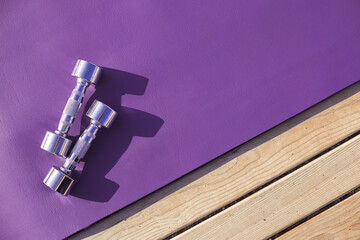 Purple yoga mat with metal chromed dumbbells. Sport fitness accessories outdoors on wooden floor on sunny day.