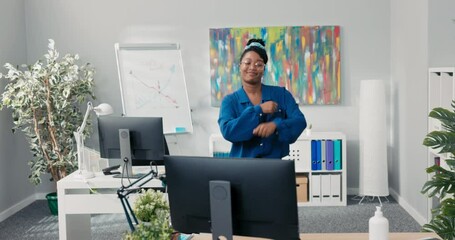 Elegant feisty girl dressed in blue shirt and stylish glasses, dancing in the middle of the office, waving her arms forward, happy for the success of the company, businesswoman with a smile on face
