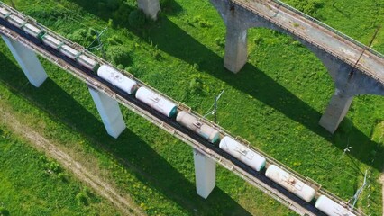 Top, aerial view of long freight train that moving along stone bridge in summer. Locomotive, tank wagons ride on rails carrying liquid or bulk cargo. Railroad transportation, delivery taken by drone