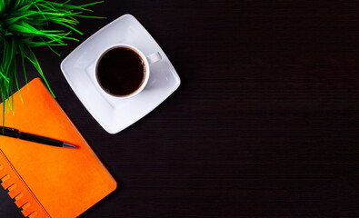 On a black background, a green plant, a white cup of coffee and an orange notepad with a pen. Business concept. Workplace close up