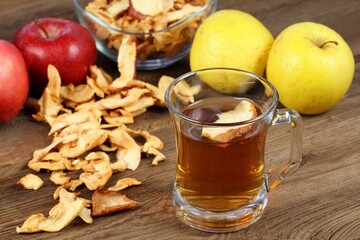 Apple tea in glass with dried slice of apple on top. Homemade tea from dried apples and hot water. Ripe red and yellow apples on brown wooden table and bowl full of dried ones.