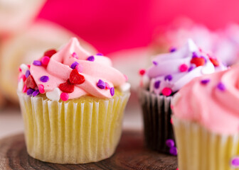 A close up view of an assortment of mini Valentine's Day cupcakes on a wooden slab.  Pink background with a very shallow depth of field.