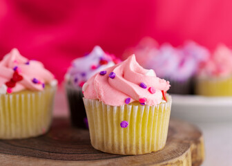 A close up view of an assortment of mini Valentine's Day cupcakes on a wooden slab.  Pink background with a very shallow depth of field.  Additional cupcakes blurred in the background.