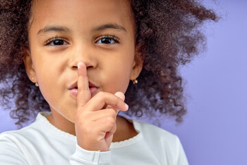 Fototapeta na wymiar Adorable black little girl holding finger on lips symbol of hush gesture of asking to be quiet. Silence or secret concept image isolated on purple studio background. close-up portrait