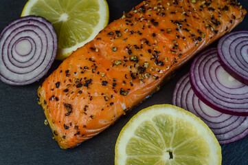 hot smoked salmon steak flavored with black pepper with lemon slices and red onion slices on a slate tray