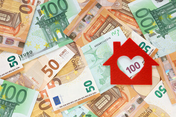 Red felt small house on right side of 50 and 100 euro banknotes background. Heart-shaped hole is cut instead of a window. Real estate loan concept. Money for a dream house. Top view. Place for text.