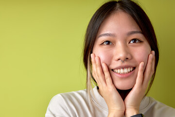 Asian women hand touch cheek looking on something with shocked and surprised face, brunette lady in casual t-shirt is smiling, in shock, posing isolated over green studio background, portrait