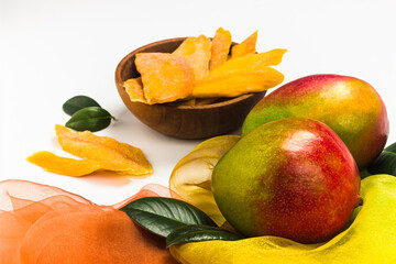 Dried mango slices in the wooden bowl, fresh mango fruits and leaves on white background. Mango products with copy space. Superfood, vegan, vegetarian food concept. Healthy snack.