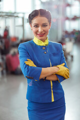 Cheerful flight attendant in air hostess uniform looking at camera and smiling while keeping arms...