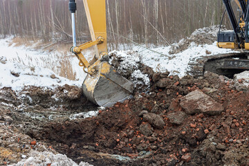 the process of loosening the soil with an excavator bucket against the background of a winter forest