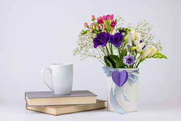 Different colors beautiful flowers bouquet in vintage white vase with ribbon and paper heart, cup of coffee or tea and books on white background. Morning concept background with copy space.