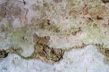 Broken wall plaster background texture with green moss. Dirty rough surface of a building facade with a big horizontal crack. Moist facade of an abandoned house exterior. Weathered condition of a wall
