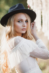 Young beautiful blond woman in white dress and dark hat posing. Attractive fashion model with loose hair