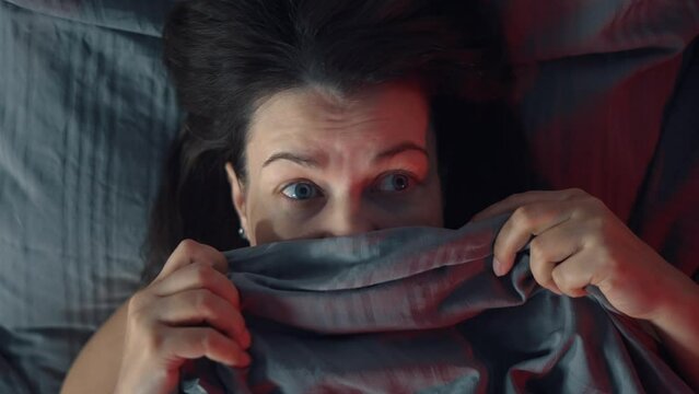 Portrait of a brunette woman in bed. She is very scared and has a grimace of horror on her face. The woman looks around and covers her face with a blanket. Nightmares and insomnia.