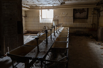 A row of old sinks in a large bathroom lit by natural window light. Abandoned public building with empty facilities. Paint is peeling off the walls because of the bad condition of the house.