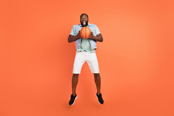 Photo of shocked guy dressed jeans waistcoat jumping high playing basketball isolated orange color background