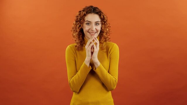 Attractive redhead caucasian girl in orange jumper twiddle her fingers and conceiving a funny plan isolated on an orange studio background.