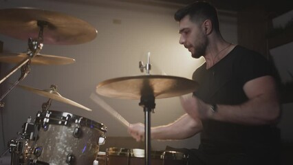 A young attractive male drummer plays with sticks on a drum kit. Makes hits on metal cymbals or Crash. Home rehearsal studio for recording sound and songs. Plays rock or heavy metal music.
