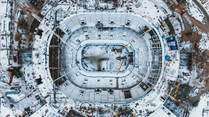 Snowy football field inside stadium with seats for spectators covered with snow and high steel building cranes pull out aerial view
