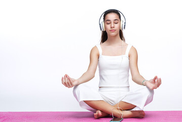 Teenage girl practicing meditation with hear eyes closed, listening to relaxing sounds on her big headphones, sitting in lotus position on a pink yoga mat