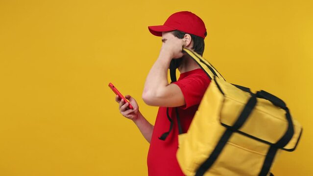 Professional delivery employee man in red cap T-shirt uniform work as dealer courier put on yellow thermal food bag backpack hold use mobile cell phone isolated plain yellow background studio portrait