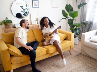 Young heterosexual couple sitting on sofa singing and playing ukulele