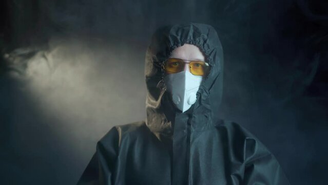 A man in a black medical suit of chemical protection and a white respirator and wearing yellow glasses and transparent medical gloves shows a gesture with his hands in the form of a cross, and then de