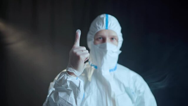 A man in a white medical suit of chemical protection and a white respirator and transparent medical gloves shows a gesture with his hands in the form of a cross, and then delivers boxing punche
