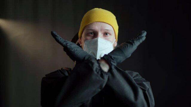A man in a black medical suit of chemical protection and a white respirator and in a yellow hat and black medical gloves makes a gesture with his hands in the form of a cross, and then shakes his head