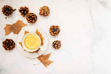 Top view of a cup of lemon tea surrounded pine cones and autumnal maple leaves. Focus on lemon.