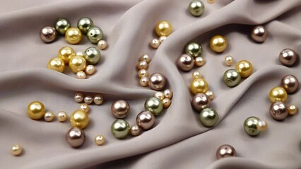 Small pink pearl beads fall on the big pearl beads different colors lying on a gray silk fabric, background, slow motion